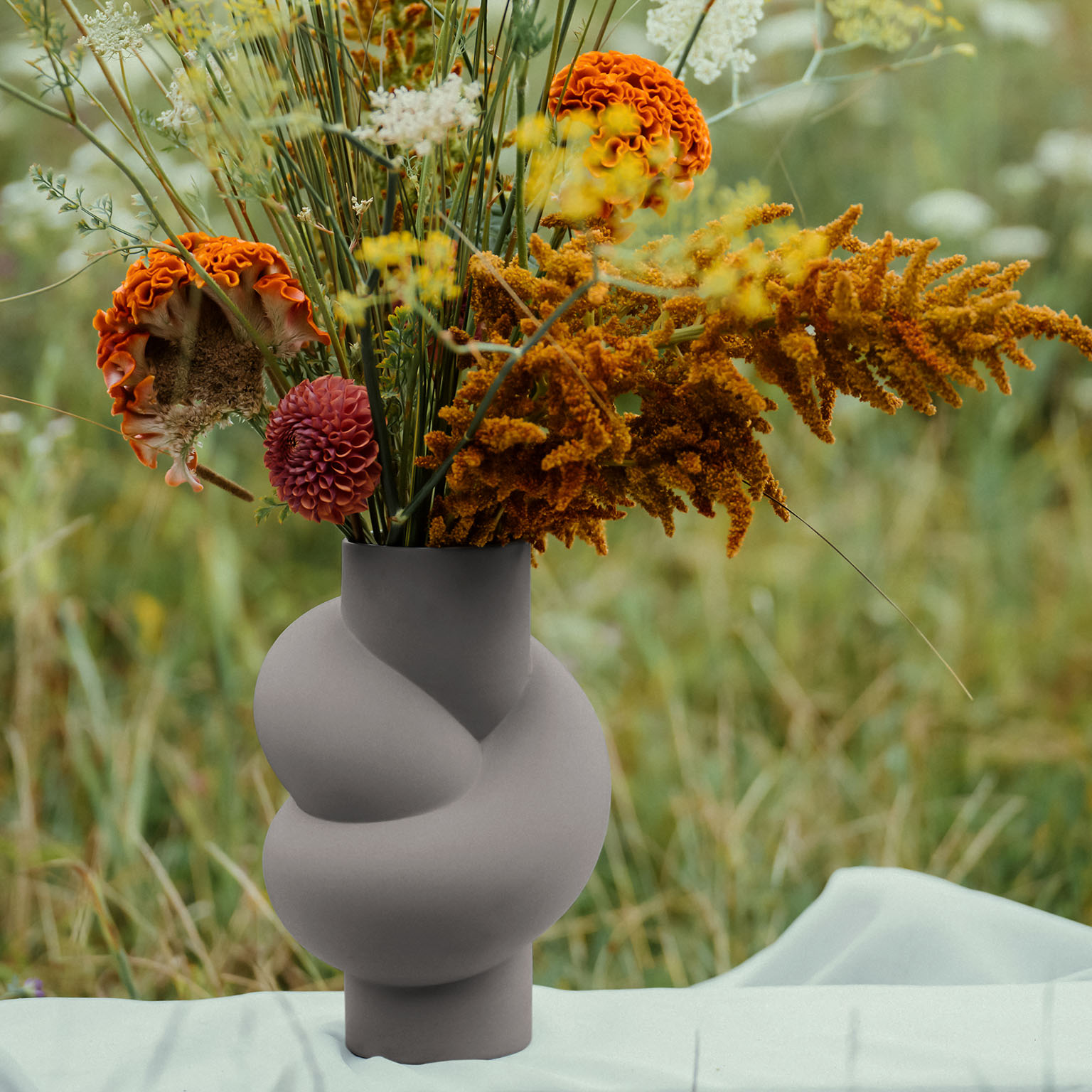 Close-up of Node Vase in Lava on a white cloth and with bouquet of flowers in orange, yellow and green.